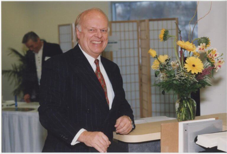 Man standing by desk smiling next to vase of flowers- obituary for Eleveld